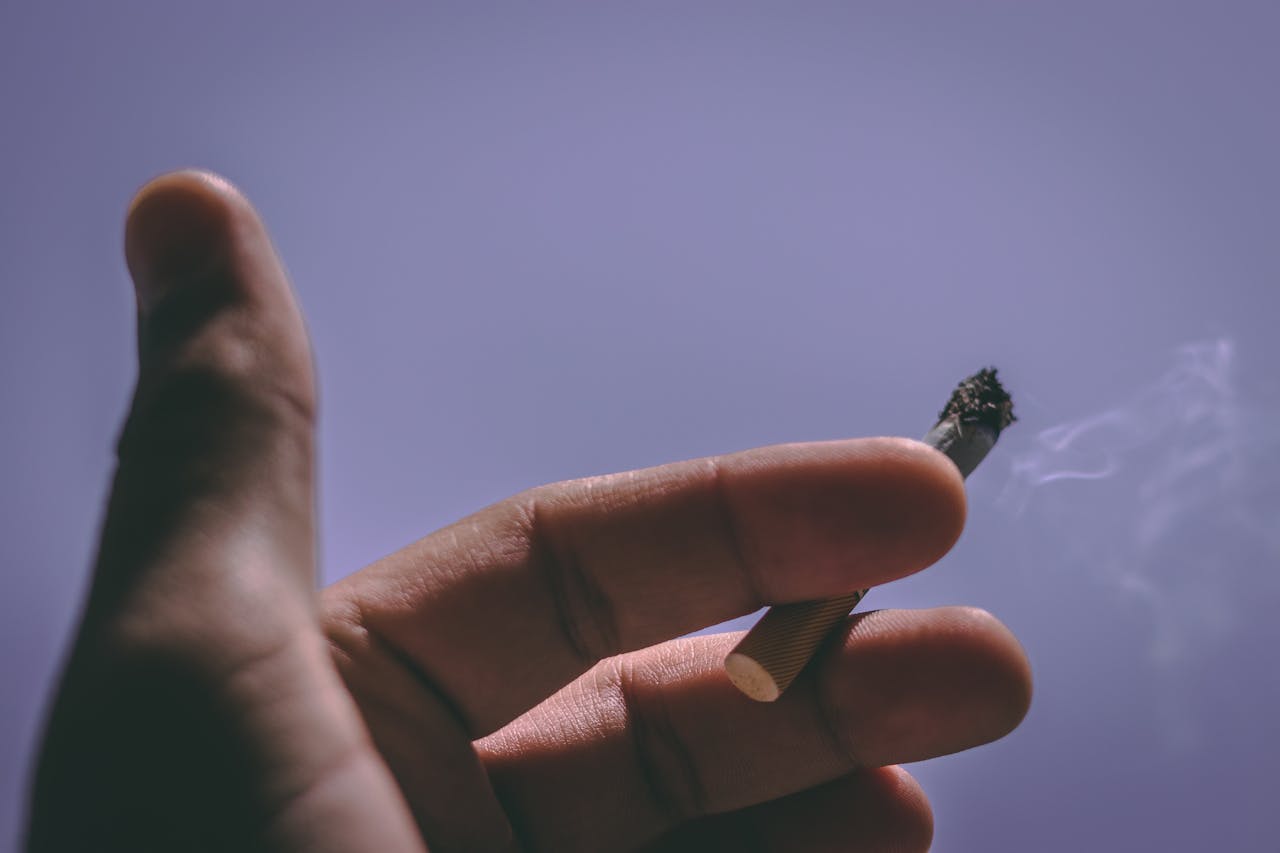 Close-up of a hand holding a lit cigarette against a clear blue sky.